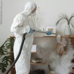 Person using fogging equipment in protective gear to disinfect indoor area with plants and bookshelves.